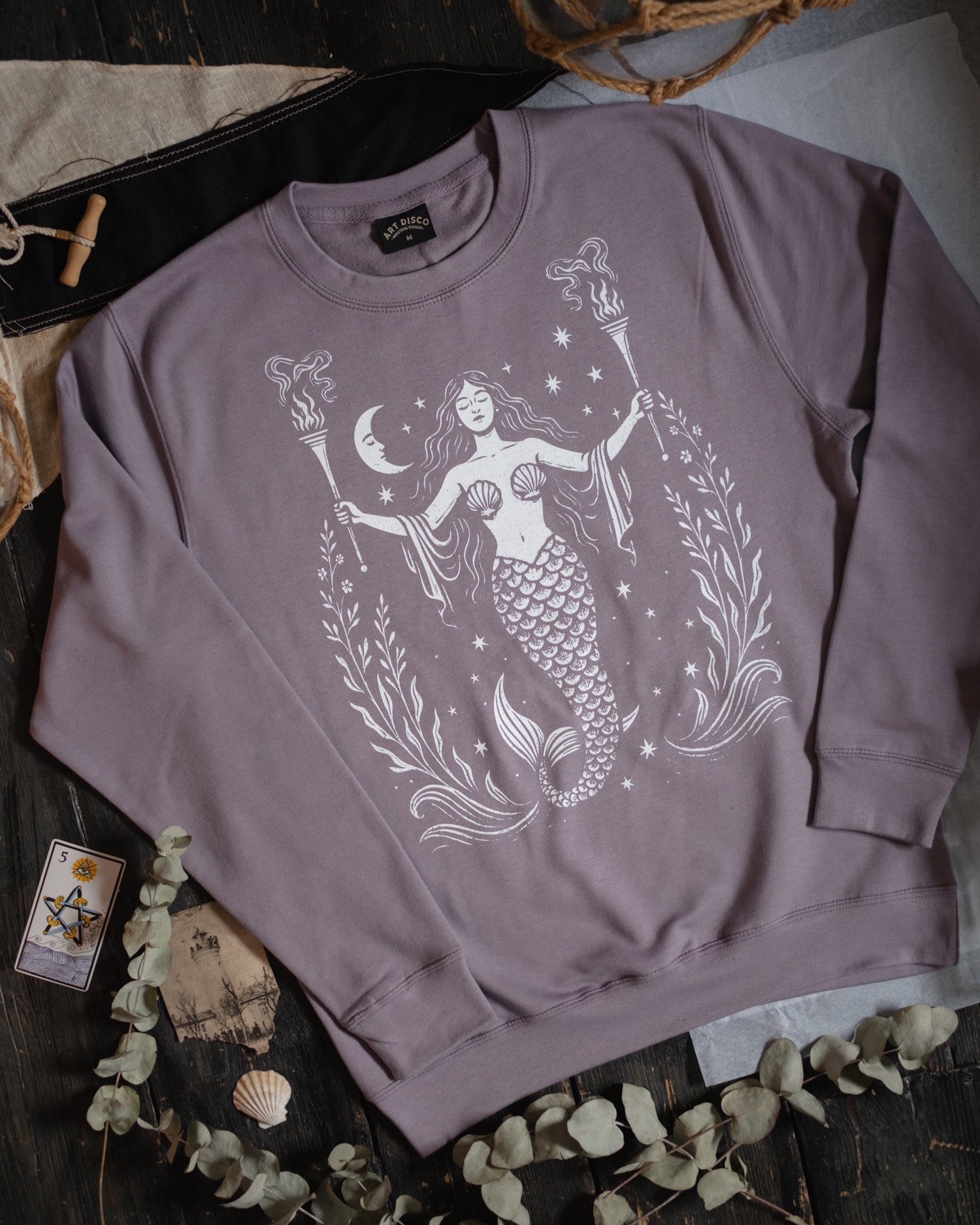 Purple Art Disco sweatshirt with mermaid design on a dark surface with decorative elements.