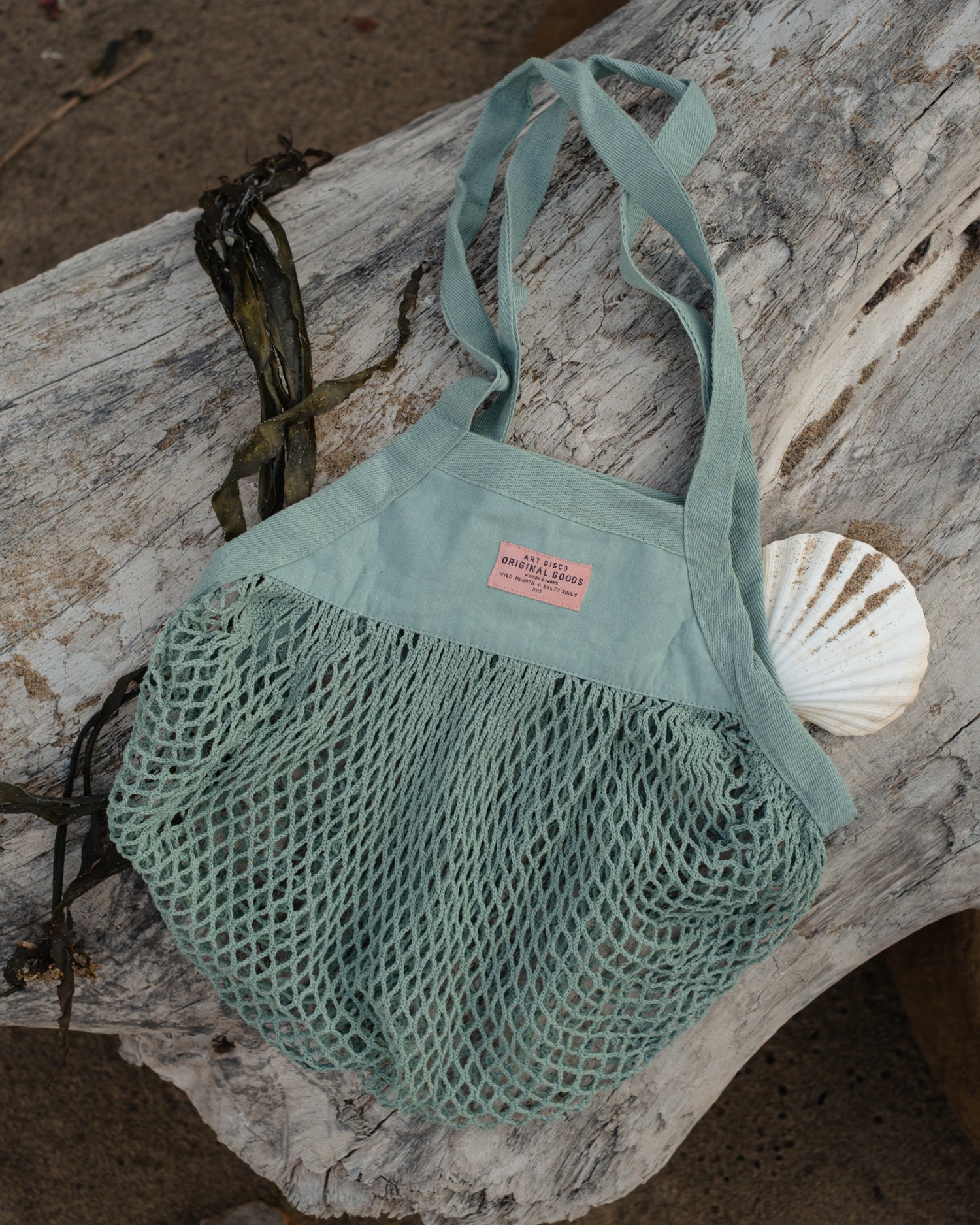 A seafoam green mesh beach tote bag with a pink brand label, styled on a piece of weathered driftwood with a seashell and seaweed.