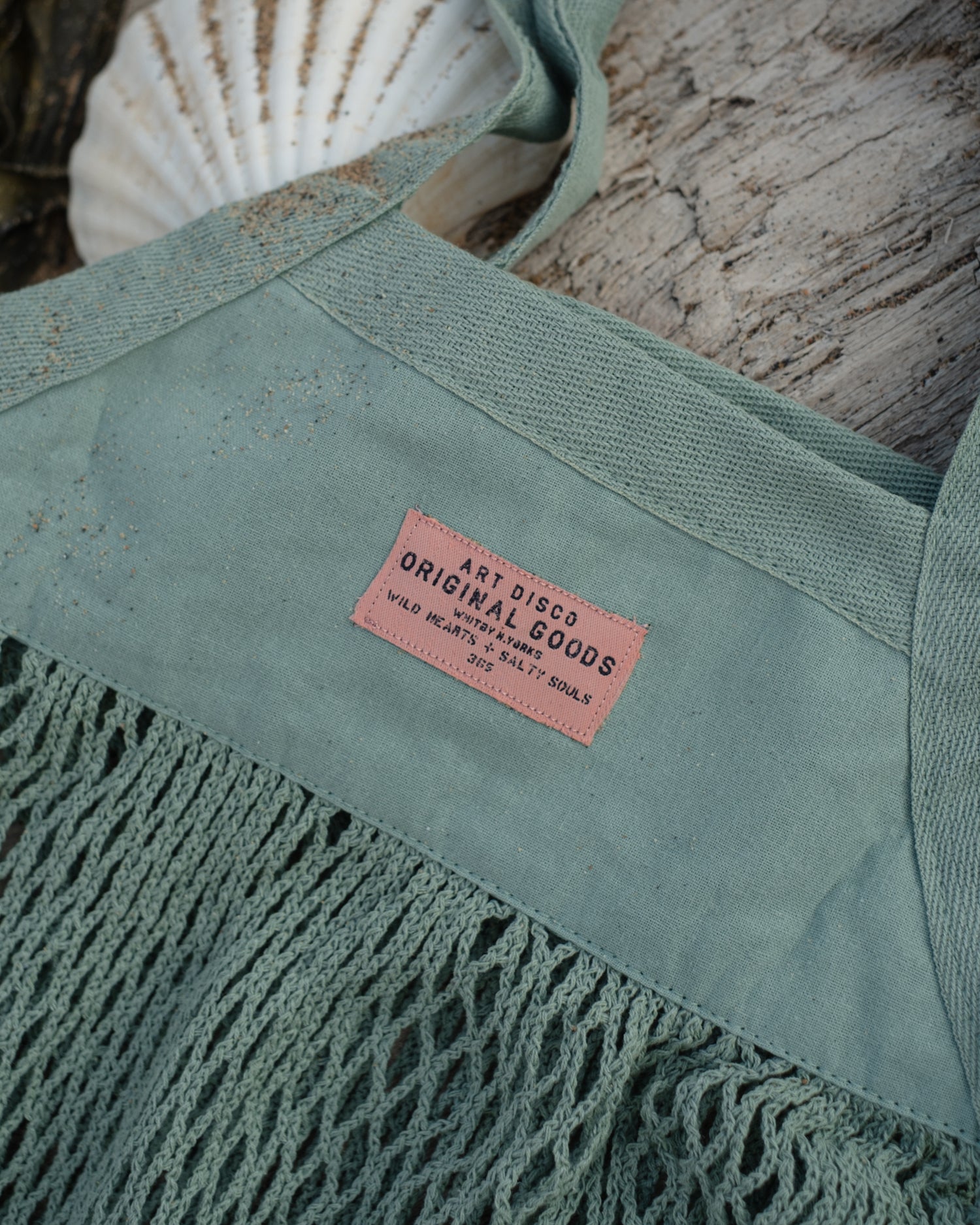 A close-up of a seafoam green mesh tote bag with a pink label reading 'ART DISCO ORIGINAL GOODS'. The bag is styled on a weathered wood surface with a white seashell in the background.