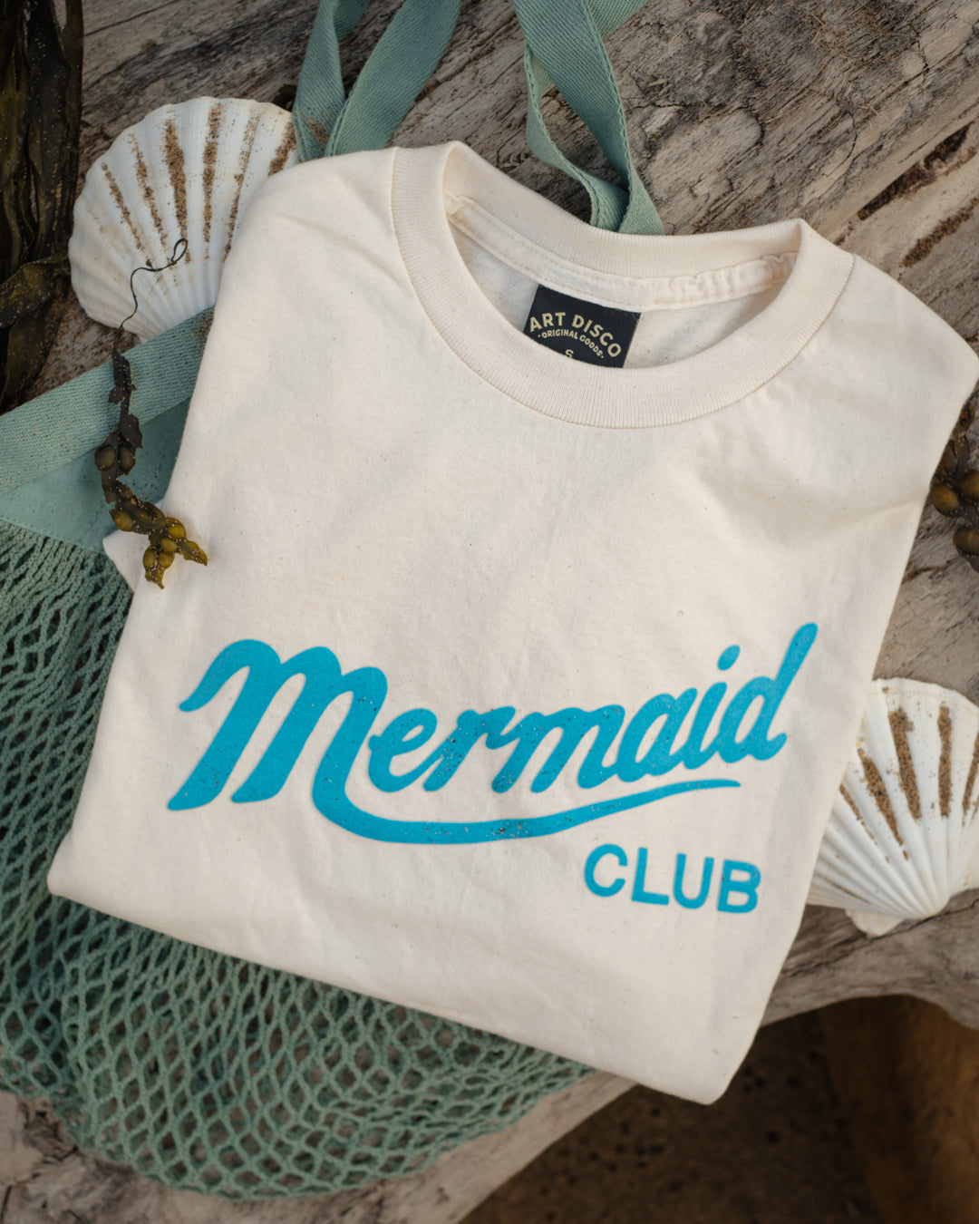 A folded off-white t-shirt with 'Mermaid CLUB' printed in turquoise script and block letters, styled on a rustic wood surface with a teal mesh bag and white seashells.