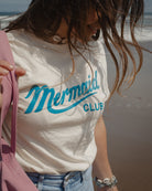 A woman on a beach wearing a cream-colored t-shirt with 'Mermaid CLUB' printed in teal script and block letters. She is also wearing light blue jeans and a pink bag is partially visible.