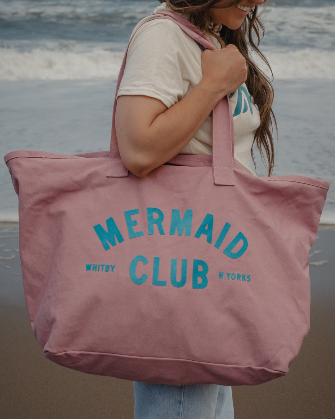 A woman on a beach carrying a large dusty pink canvas tote bag with 'MERMAID CLUB' printed in teal. The background shows the ocean waves and sandy shore.