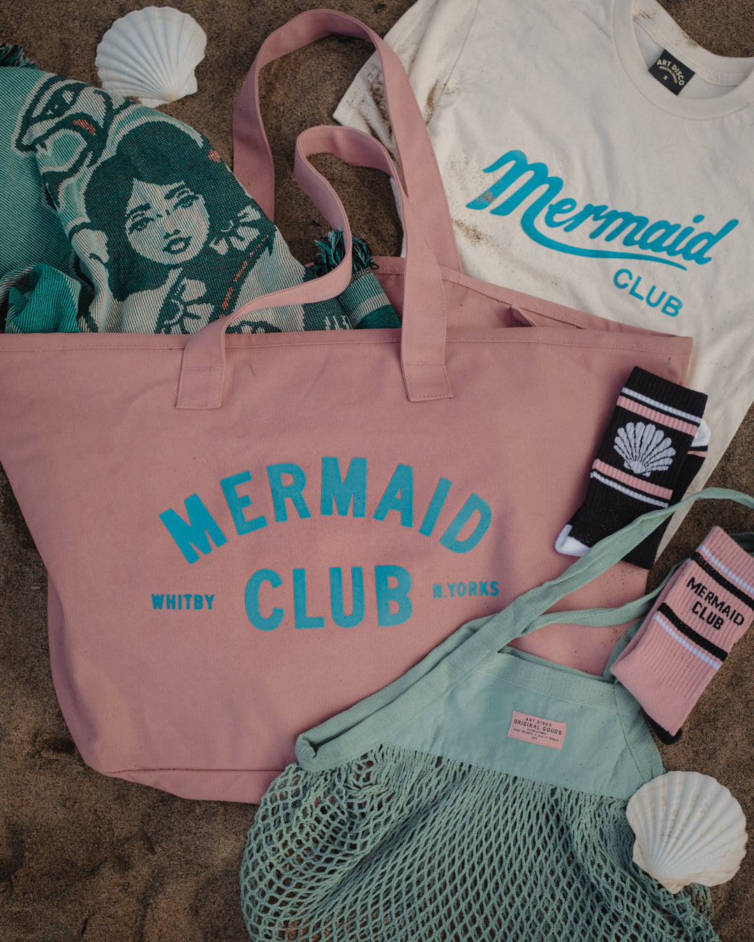 A flat-lay arrangement of a pink canvas tote bag with 'MERMAID CLUB' printed in teal, alongside a mint green mesh bag, a white t-shirt, socks, and a patterned blanket on a sandy surface with seashell props.