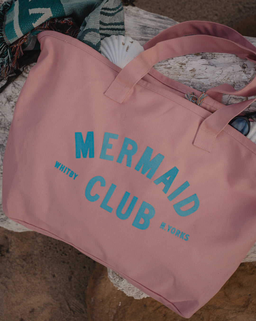 A dusty pink canvas tote bag with 'MERMAID CLUB, WHITBY N.YORKS' printed in light blue, styled on a rustic wooden surface with a striped knit blanket and a seashell nearby.