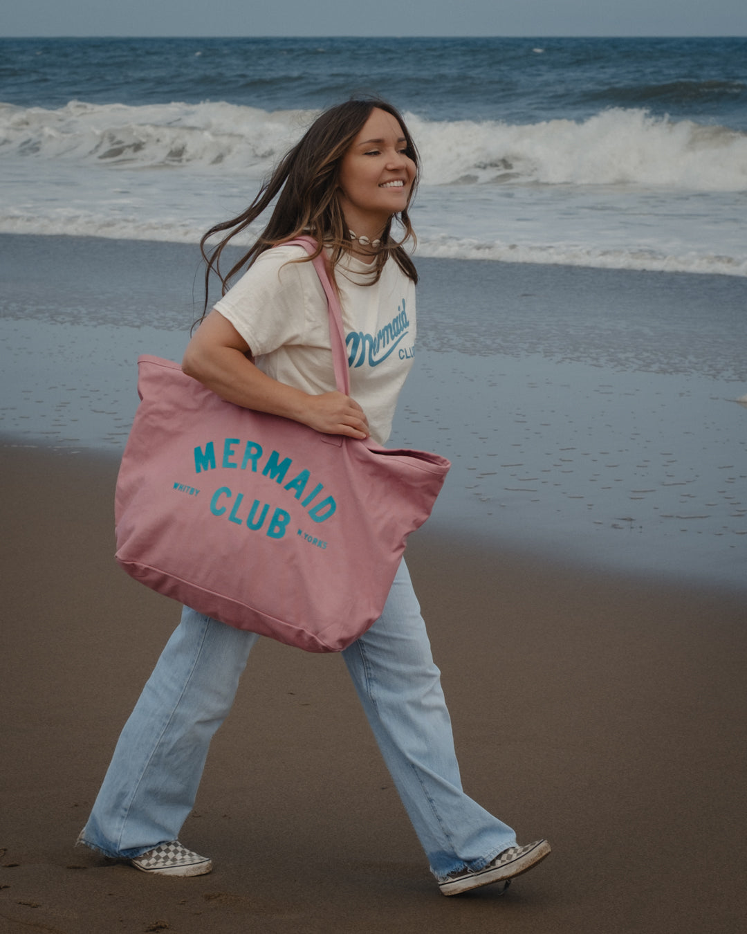 A woman walking on a beach carrying a large dusty pink canvas tote bag with 'Mermaid Club' printed in blue. She is wearing a cream graphic t-shirt, light blue jeans, and slip-on shoes.