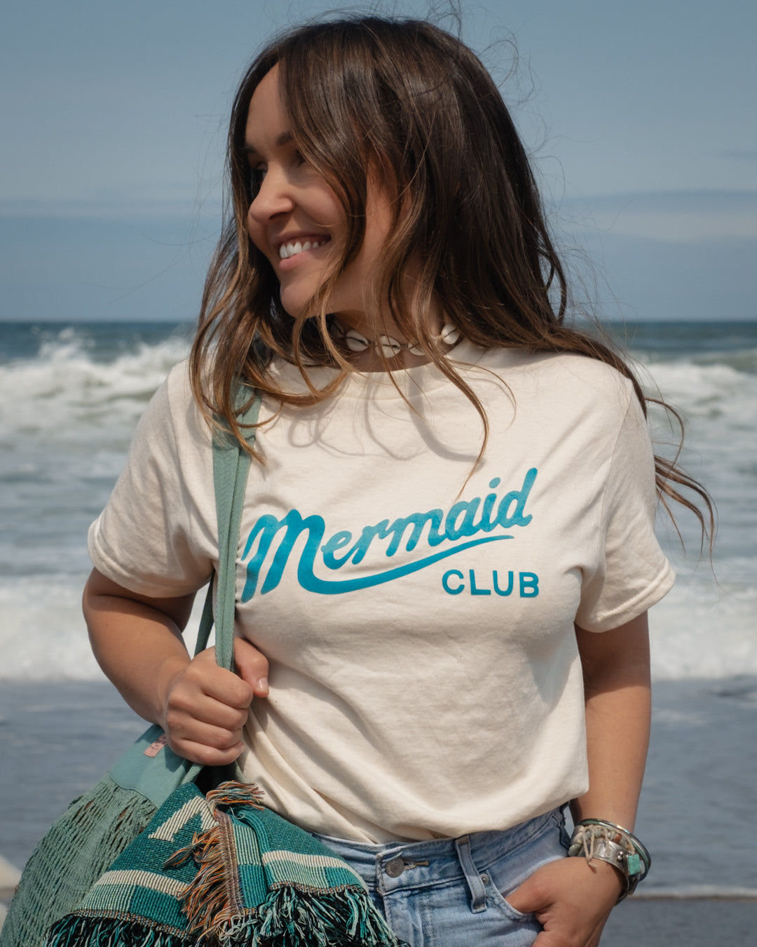 A woman standing on a beach wearing a cream-colored t-shirt with 'Mermaid CLUB' printed in turquoise, paired with light blue jeans and carrying a teal bag & blanket.