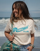 A woman standing on a beach wearing a cream-colored t-shirt with 'Mermaid CLUB' printed in turquoise, paired with light blue jeans and carrying a teal bag & blanket.