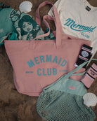 A flat-lay arrangement of a pink canvas tote bag with 'MERMAID CLUB' printed in teal, alongside a mint green mesh bag, a white t-shirt, socks, and a patterned blanket on a sandy surface with seashell props.