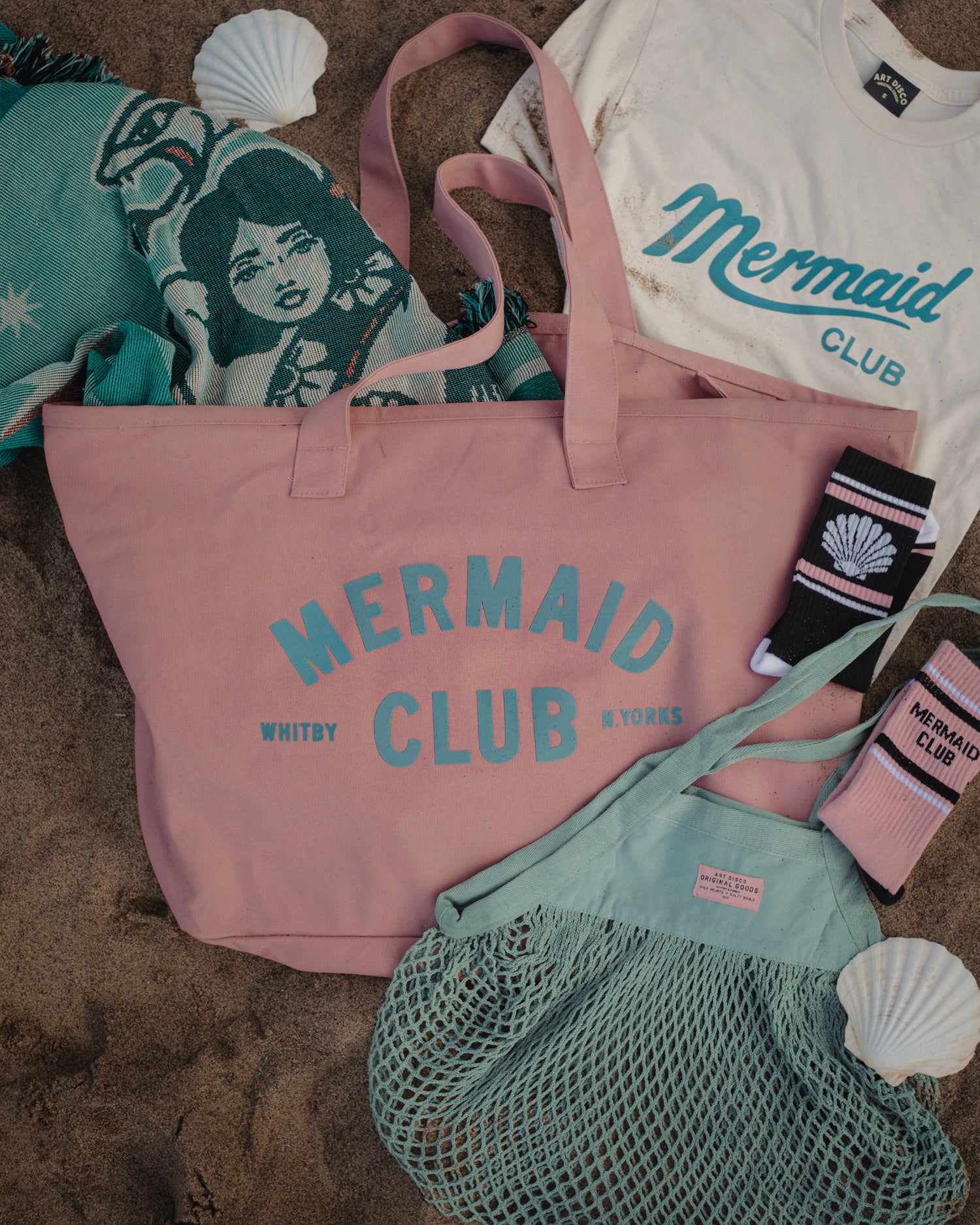 A flat-lay arrangement of a pink canvas tote bag with 'MERMAID CLUB' printed in teal, alongside a mint green mesh bag, a white t-shirt, socks, and a patterned blanket on a sandy surface with seashell props.