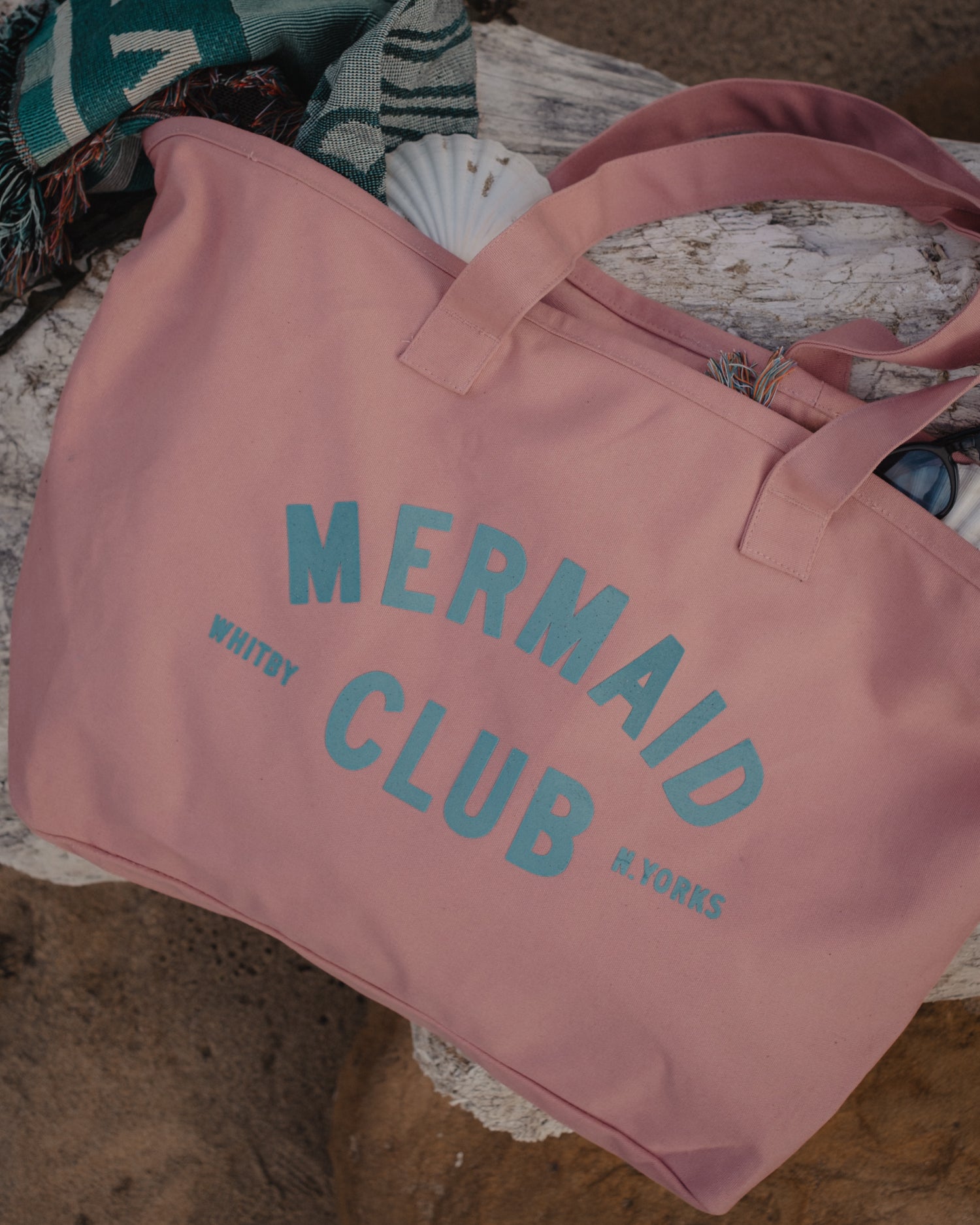A dusty pink canvas tote bag with 'MERMAID CLUB, WHITBY N.YORKS' printed in light blue, styled on a rustic wooden surface with a striped knit blanket and a seashell nearby.