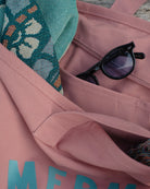 A dusty pink canvas tote bag with a zipper pocket, shown in a close-up flat-lay style. A pair of black sunglasses rests inside the pocket, and a teal patterned textile is draped over the top edge.