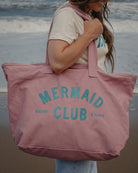 A woman on a beach carrying a large dusty pink canvas tote bag with 'MERMAID CLUB' printed in teal. The background shows the ocean waves and sandy shore.