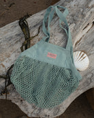 A seafoam green mesh beach tote bag with a pink brand label, styled on a piece of weathered driftwood with a seashell and seaweed.
