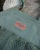 A close-up of a seafoam green mesh tote bag with a pink label reading 'ART DISCO ORIGINAL GOODS'. The bag is styled on a weathered wood surface with a white seashell in the background.