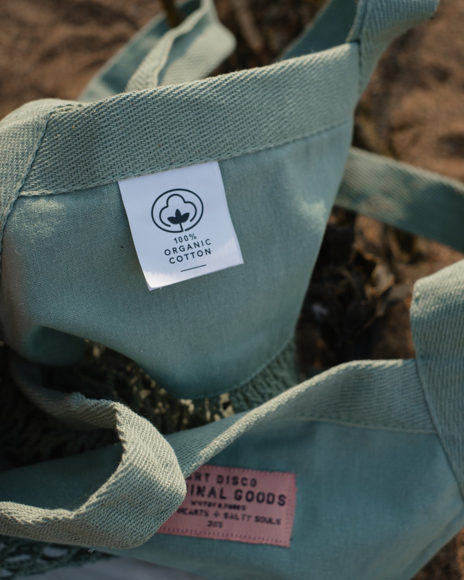 A close-up of a seafoam green mesh shopper tote bag with a white '100% Organic Cotton' label and a pink brand tag, resting on sand.