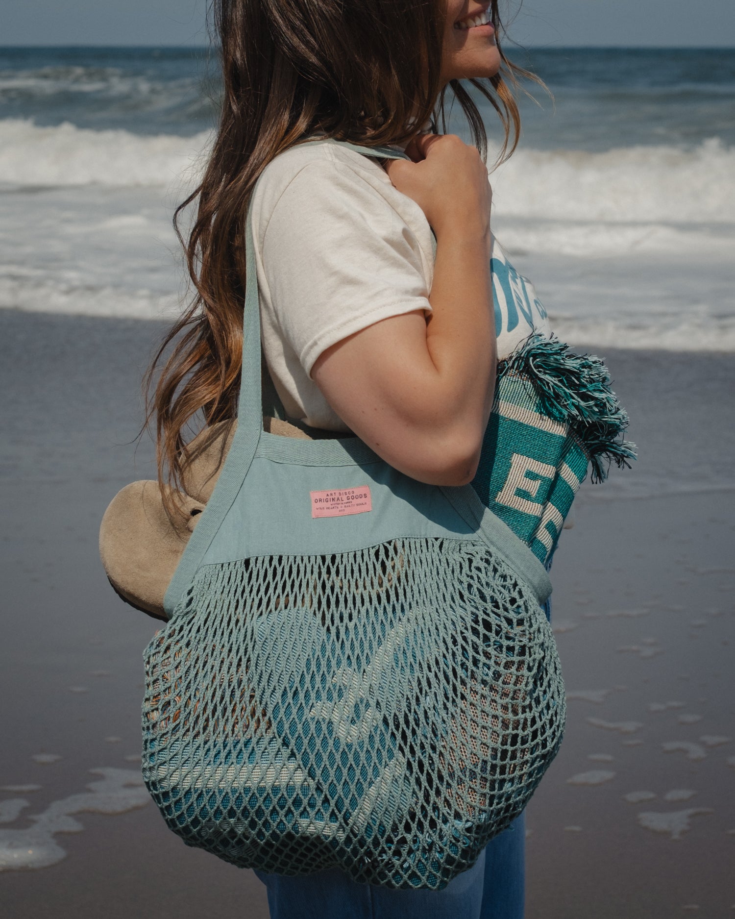 A woman on a beach wearing a seafoam green mesh tote bag with a pink label. The bag contains a pair of shoes and a teal knit blanket with tassels.