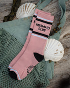 A pair of pink crew socks with black and white stripes and the words 'A MERMAID CLUB' on the cuff, styled on a green mesh bag with seashells and wood.