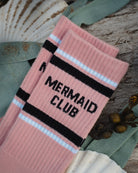 A pair of pink ribbed crew socks with black and white stripes and the words 'MERMAID CLUB' knitted on the side, styled on a rustic wood surface with seashells and green fabric.