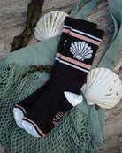 A pair of black crew sock with pink and white stripes, a white scallop shell graphic, and white toe/heel sections, styled on a green mesh bag with seashells and wood background.
