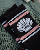 A pair of black ribbed socks with a white seashell motif and pink and white stripes, styled on a textured green knit blanket.