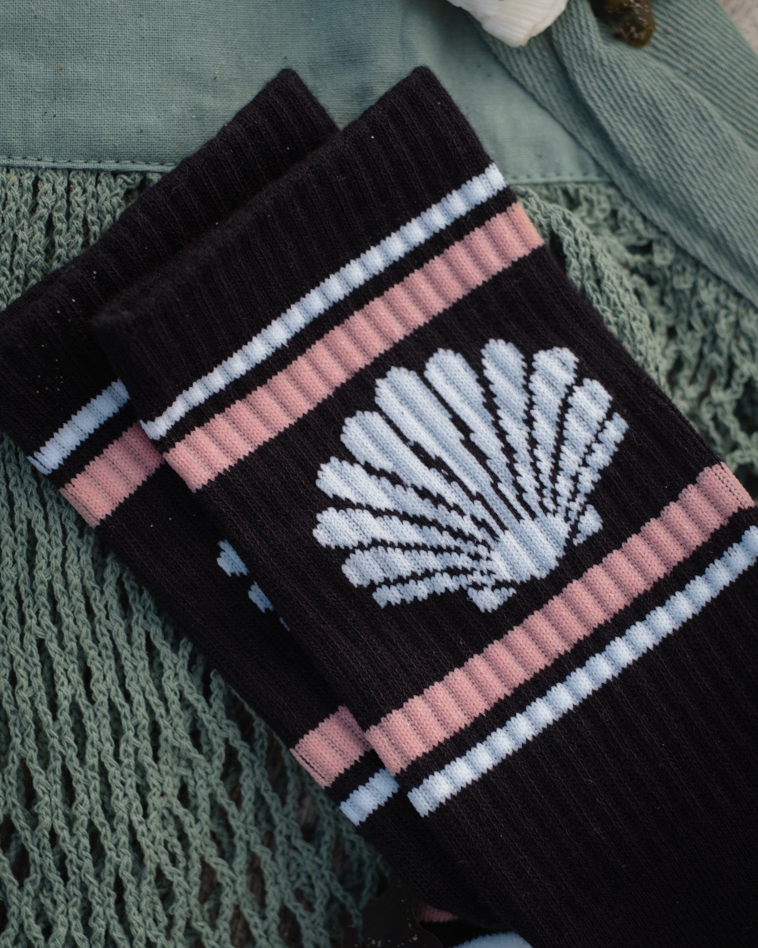 A pair of black ribbed socks with a white seashell motif and pink and white stripes, styled on a textured green knit blanket.
