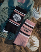 Two crew socks styled on a sandy beach with seashells and a green net. One sock is black with pink stripes, a white seashell graphic, and white text.