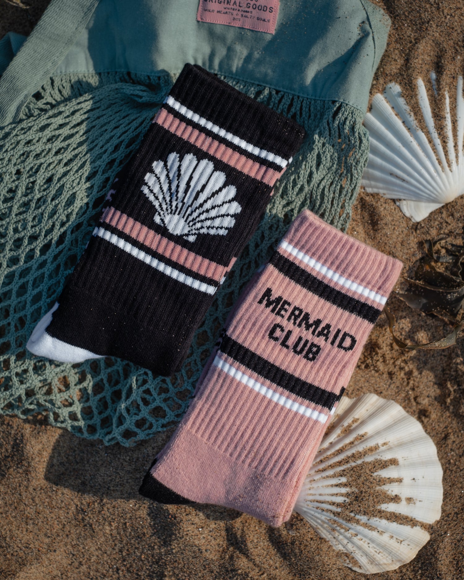 Two crew socks styled on a sandy beach with seashells and a green net. One sock is black with pink stripes, a white seashell graphic, and white text.