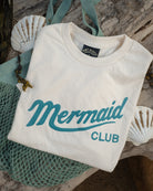 A folded off-white t-shirt with 'Mermaid CLUB' printed in teal script and block letters, styled on a rustic wood surface with a teal mesh bag and white seashells.