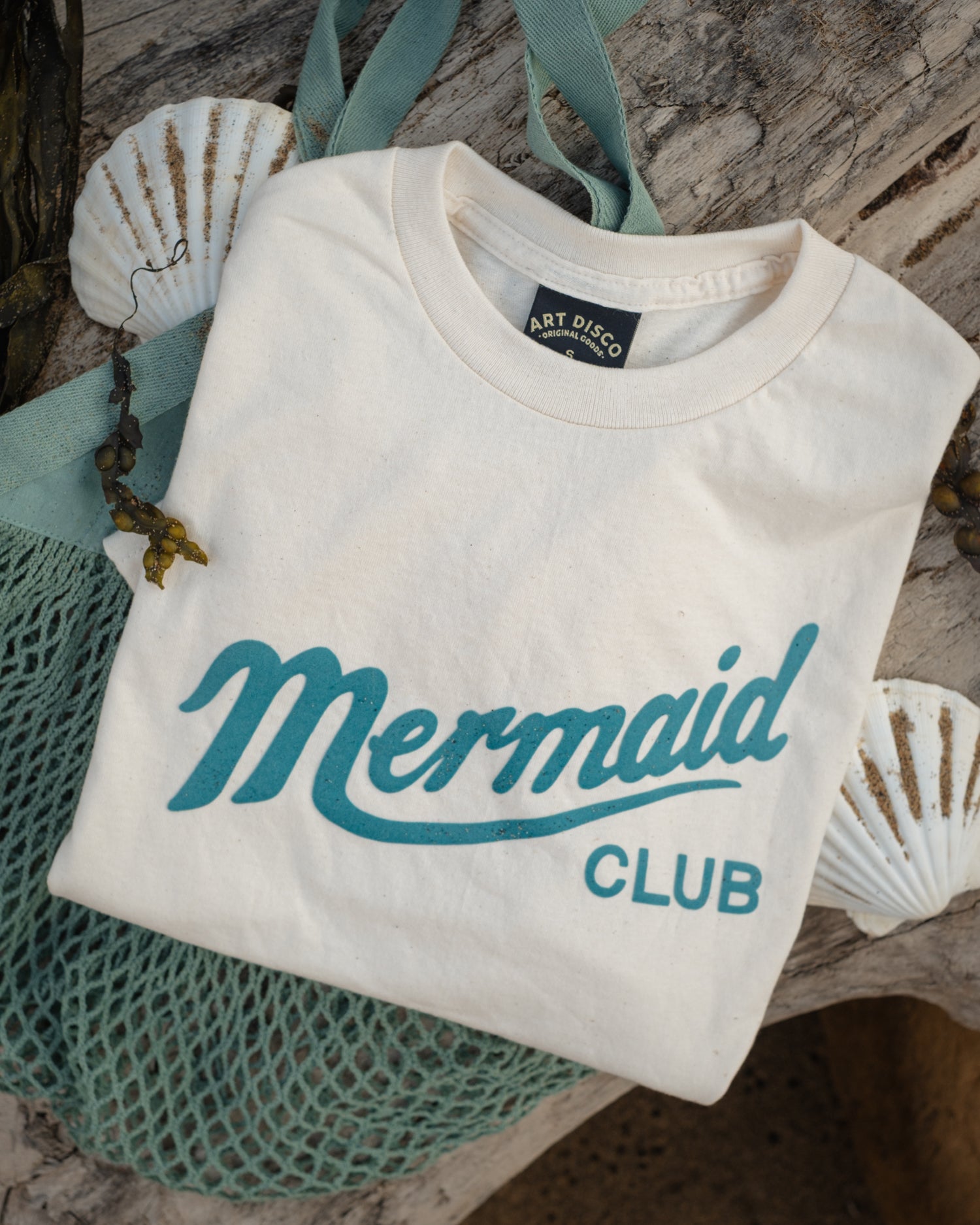 A folded off-white t-shirt with 'Mermaid CLUB' printed in teal script and block letters, styled on a rustic wood surface with a teal mesh bag and white seashells.