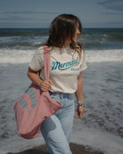 A woman walking on a beach wearing a cream t-shirt with 'Mermaid CLUB' printed in teal, paired with light blue jeans and carrying a dusty pink tote bag with the same text.