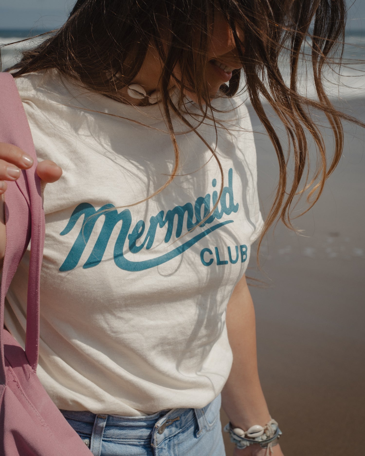 A woman on a beach wearing a cream-colored t-shirt with 'Mermaid CLUB' printed in teal script and block letters. She is also wearing light blue jeans and a pink bag is partially visible.
