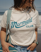A woman wearing a cream-colored t-shirt with 'Mermaid CLUB' printed in teal across the chest, photographed outdoors at the beach with ocean waves in the background.