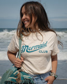 A woman standing on a beach wearing a cream-colored t-shirt with 'Mermaid CLUB' printed in teal, paired with light blue jeans and carrying a teal bag & blanket.