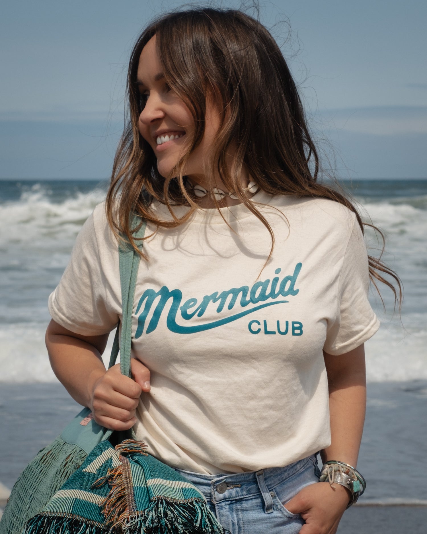 A woman standing on a beach wearing a cream-colored t-shirt with 'Mermaid CLUB' printed in teal, paired with light blue jeans and carrying a teal bag & blanket.